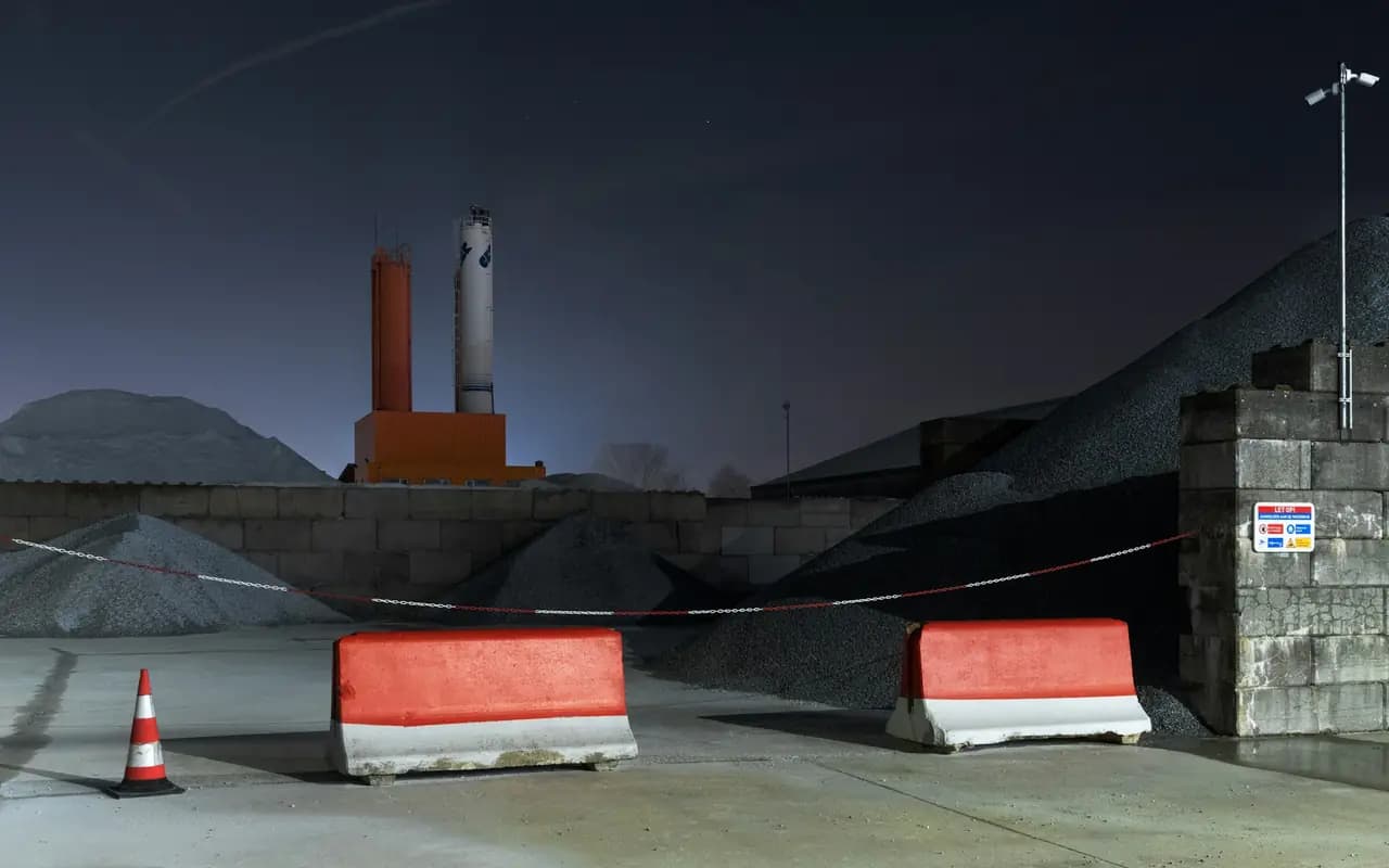 Cour d’un dépôt de matériaux la nuit, déserte : tas de granulats, séparateurs de chantier rouges et blancs et cône de signalisation sous l’éclairage du site.