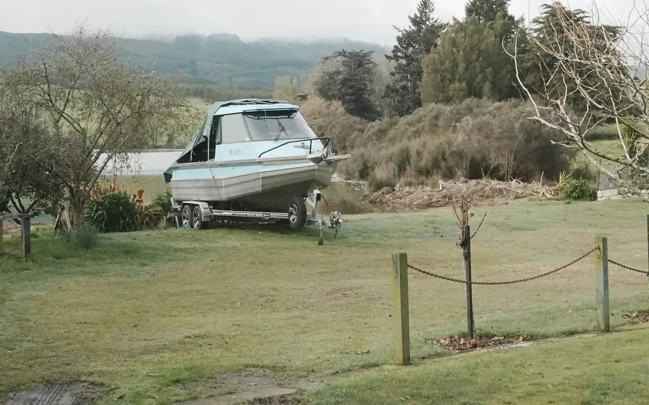 Vedette à cabine posée sur sa remorque routière, garée sur l’herbe d’un terrain bordé d’une clôture, au bord d’un plan d’eau.