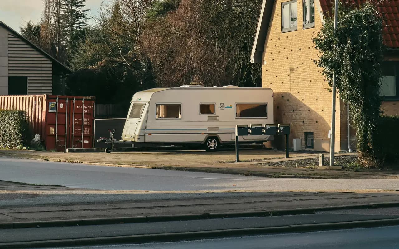 Caravane de tourisme dételée et remisée sur un terrain gravillonné à côté d’une maison, arbres nus et lumière d’hiver.