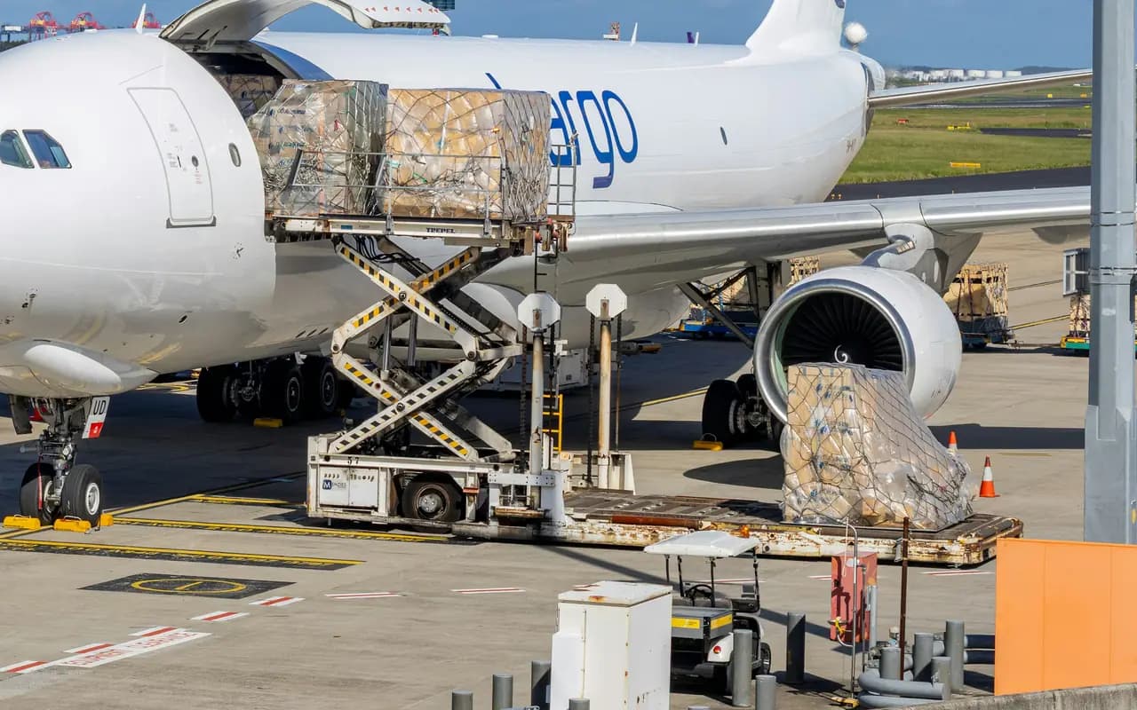 Porte cargo latérale d’un avion-cargo ouverte au poste de chargement, palettes filmées présentées par un élévateur à ciseaux sur le tarmac.