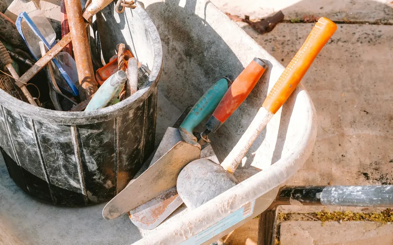 Seau de chantier rempli de truelles et d’outils de maçon, posé contre une brouette sur une terrasse en dalles en cours de pose.
