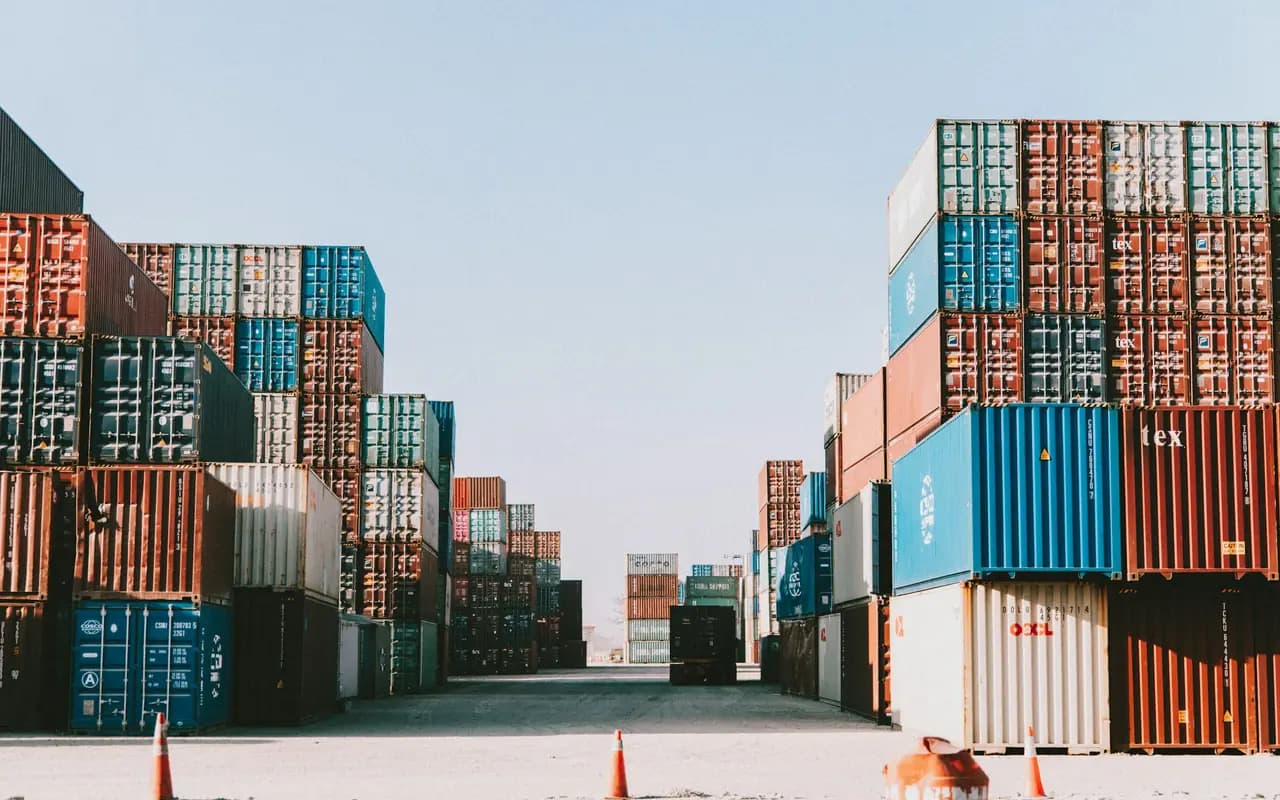 Allée déserte entre deux piles de conteneurs maritimes empilés sur quatre hauteurs, dans un parc de stockage au sol sec.